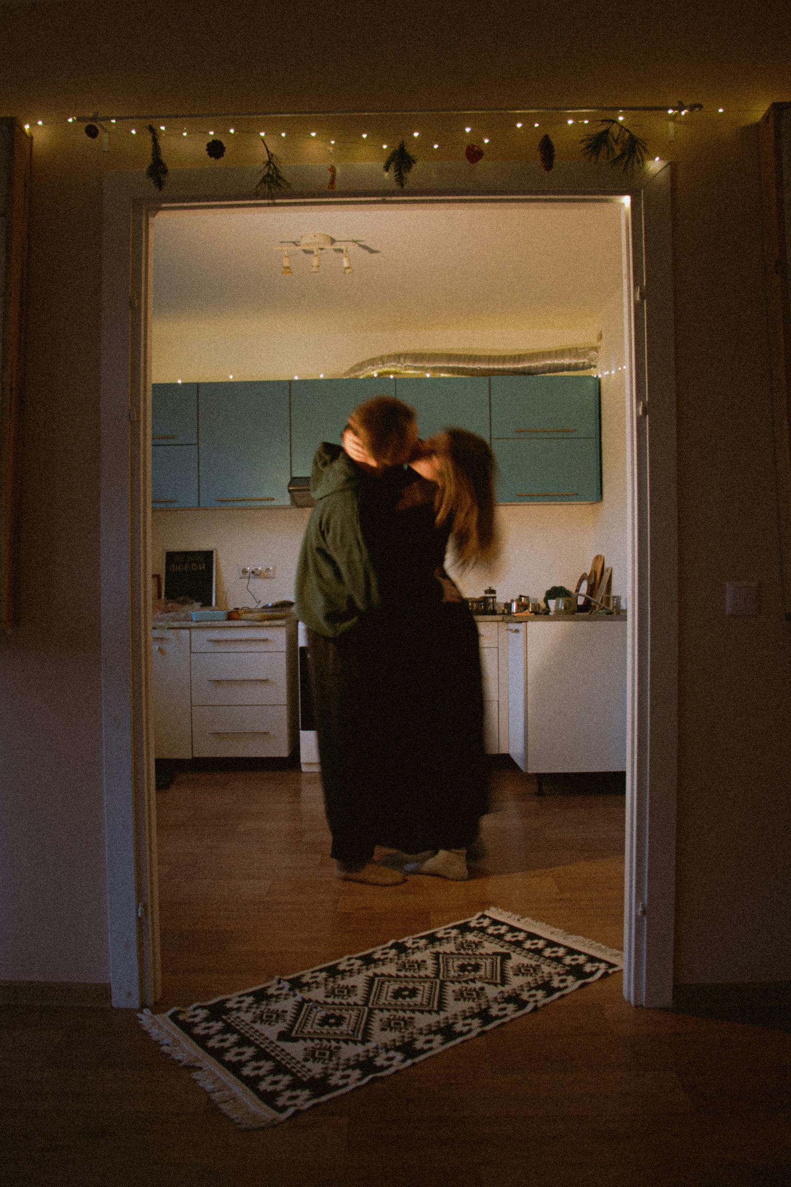 A couple embracing in their kitchen doorway, lit by string lights.